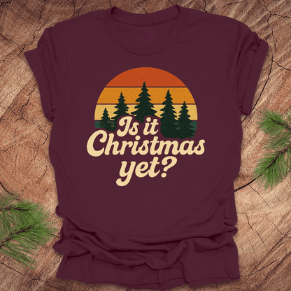 Maroon t-shirt with 'Is it Christmas yet?' graphic on a wooden surface with pine branches.