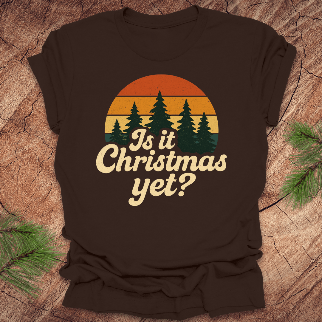 Brown t-shirt with 'Is it Christmas yet?' graphic on a wooden surface with pine branches.