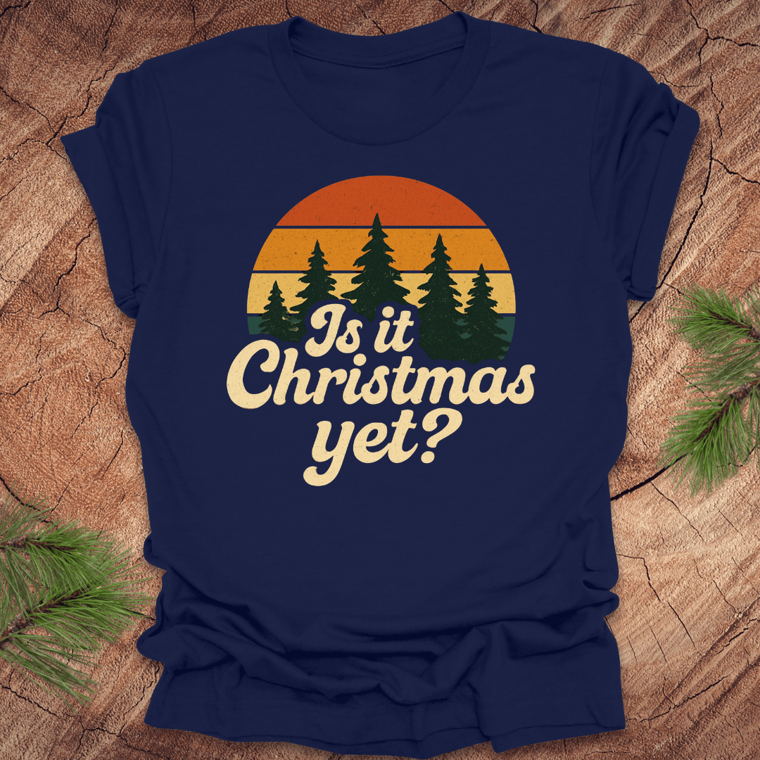 Navy blue t-shirt with 'Is it Christmas yet?' design on a wooden surface with pine branches.