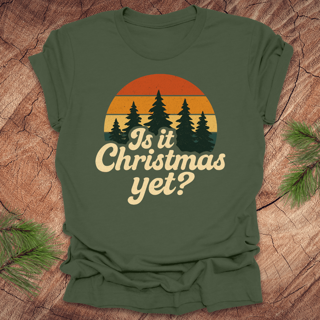 Green t-shirt with 'Is it Christmas yet?' design on a wooden surface with pine branches.