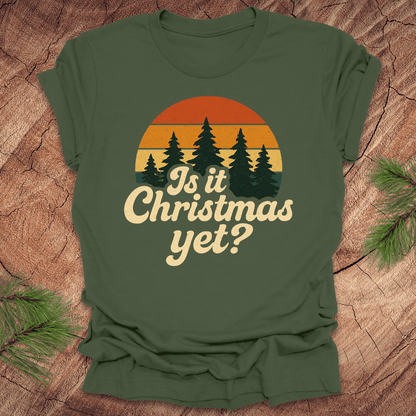 Green t-shirt with 'Is it Christmas yet?' design on a wooden surface with pine branches.