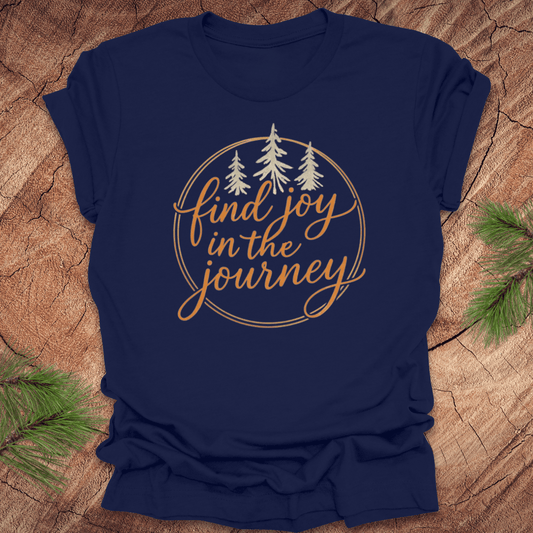 Navy blue t-shirt with 'find joy in the journey' text on a wooden surface with pine branches.