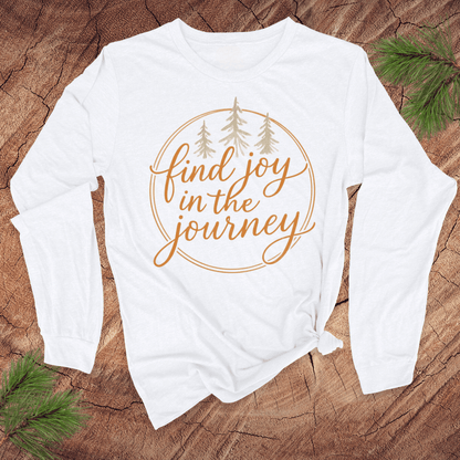 White long-sleeve shirt with 'find joy in the journey' text on a wooden surface with pine branches.