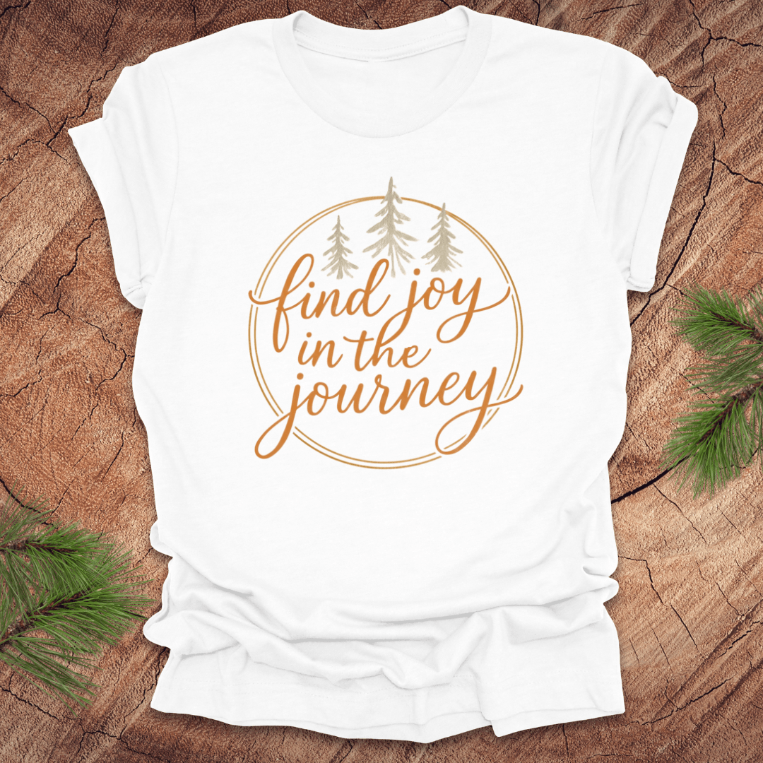 White t-shirt with 'find joy in the journey' text on a wooden surface with pine branches.