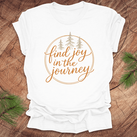White t-shirt with 'find joy in the journey' text on a wooden surface with pine branches.