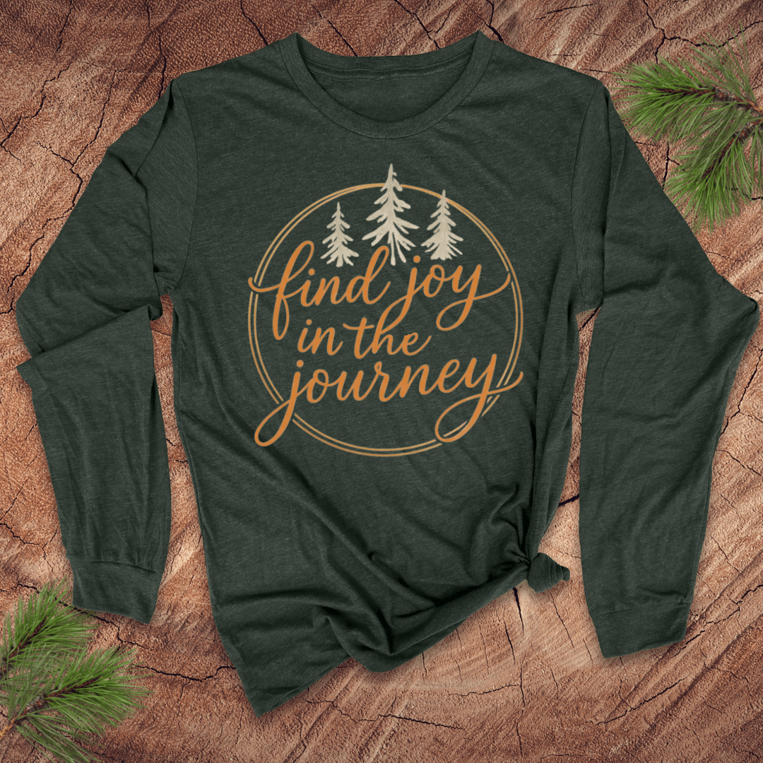 Green long-sleeve shirt with 'find joy in the journey' text on a wooden surface with pine branches.