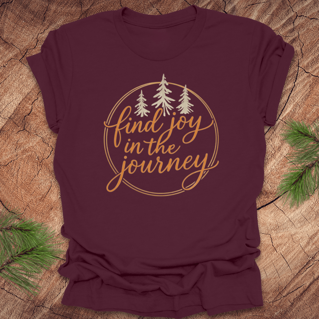 Maroon t-shirt with 'find joy in the journey' text and trees on a wooden surface.