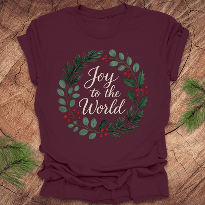 Maroon t-shirt with wreath and text, 'Joy to the World' design on a wooden surface