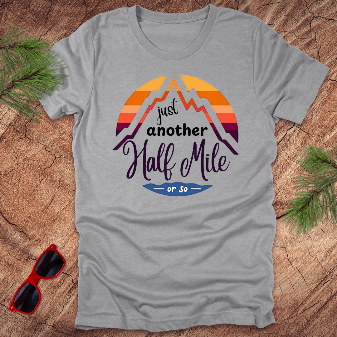 A gray t-shirt with a mountain graphic and the text 'Just Another Half Mile' on the front, displayed on a wooden surface with pine needles and a pair of sunglasses in the background.