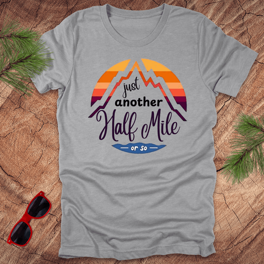 A gray t-shirt with a mountain graphic and the text 'Just Another Half Mile' on the front, displayed on a wooden surface with pine needles and a pair of sunglasses in the background.