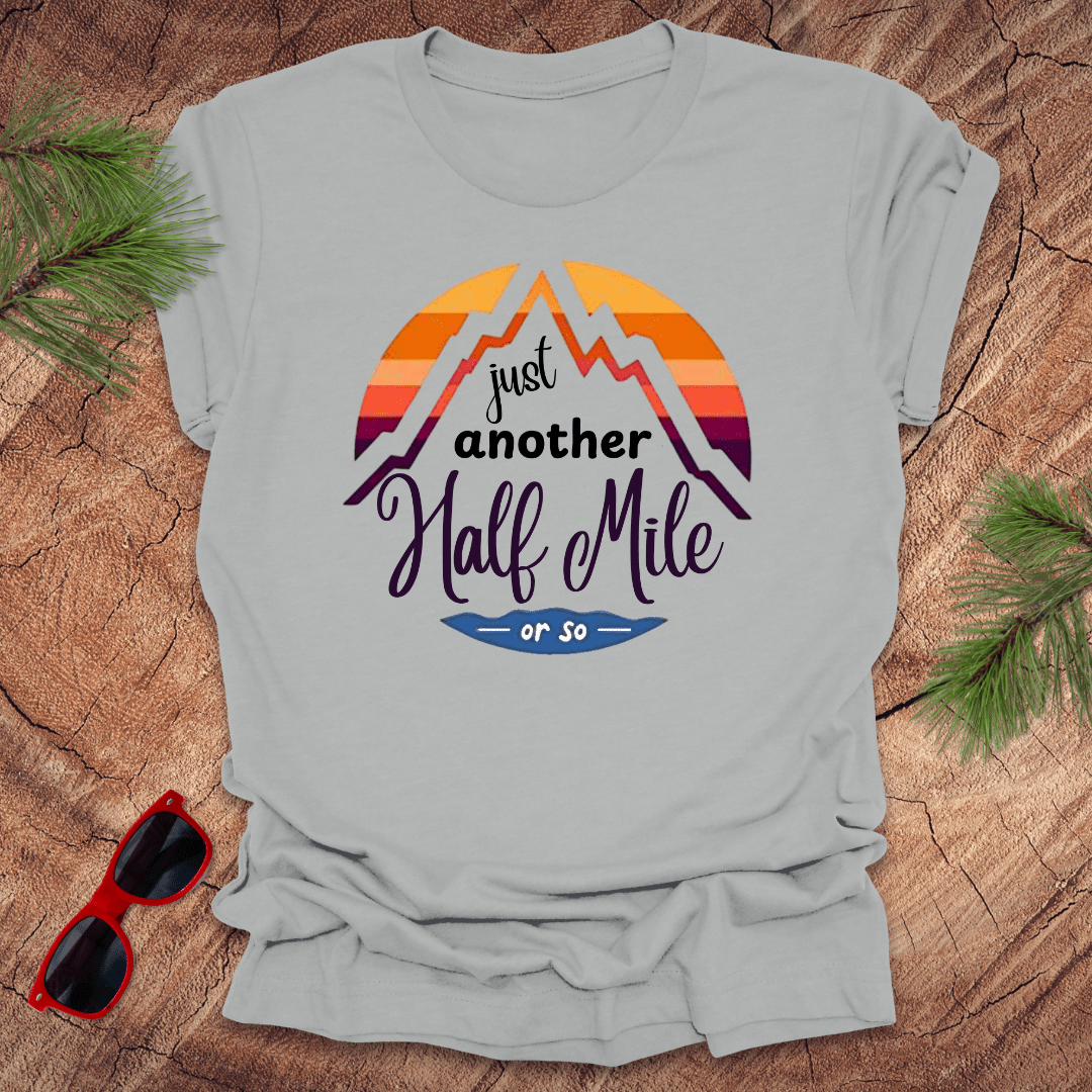 A silver t-shirt with a mountain graphic and the text 'Just Another Half Mile' on the front, displayed on a wooden surface with pine needles and a pair of sunglasses in the background.