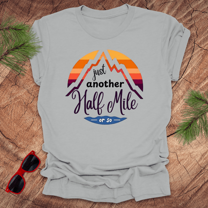A silver t-shirt with a mountain graphic and the text 'Just Another Half Mile' on the front, displayed on a wooden surface with pine needles and a pair of sunglasses in the background.
