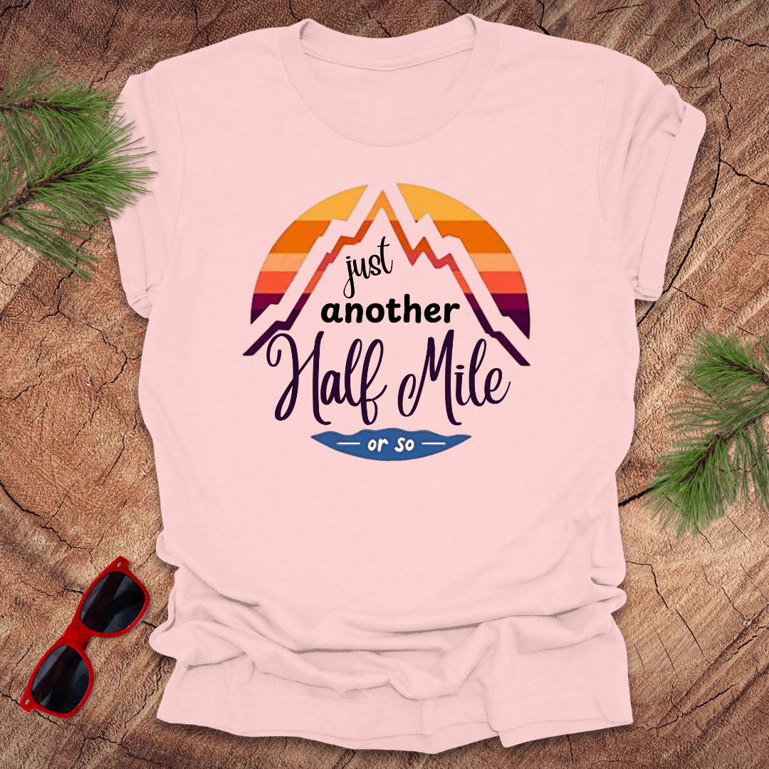 A pink t-shirt with a mountain graphic and the text 'Just Another Half Mile' on the front, displayed on a wooden surface with pine needles and a pair of sunglasses in the background.