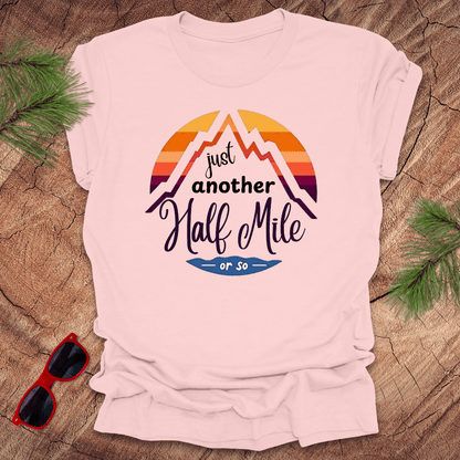 A pink t-shirt with a mountain graphic and the text 'Just Another Half Mile' on the front, displayed on a wooden surface with pine needles and a pair of sunglasses in the background.