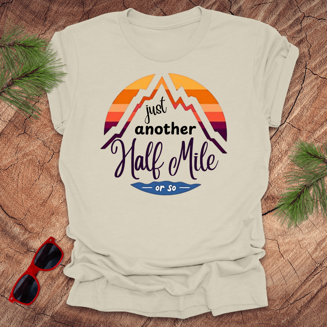 A natural t-shirt with a mountain graphic and the text 'Just Another Half Mile' on the front, displayed on a wooden surface with pine needles and a pair of sunglasses in the background.