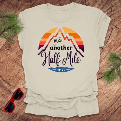 A natural t-shirt with a mountain graphic and the text 'Just Another Half Mile' on the front, displayed on a wooden surface with pine needles and a pair of sunglasses in the background.