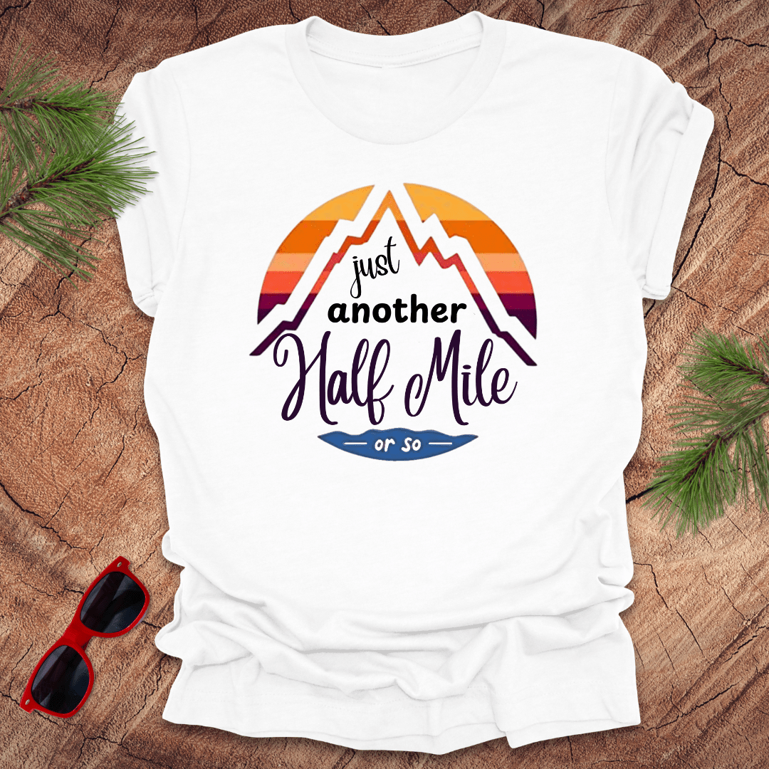 A white t-shirt with a mountain graphic and the text 'Just Another Half Mile' on the front, displayed on a wooden surface with pine needles and a pair of sunglasses in the background.