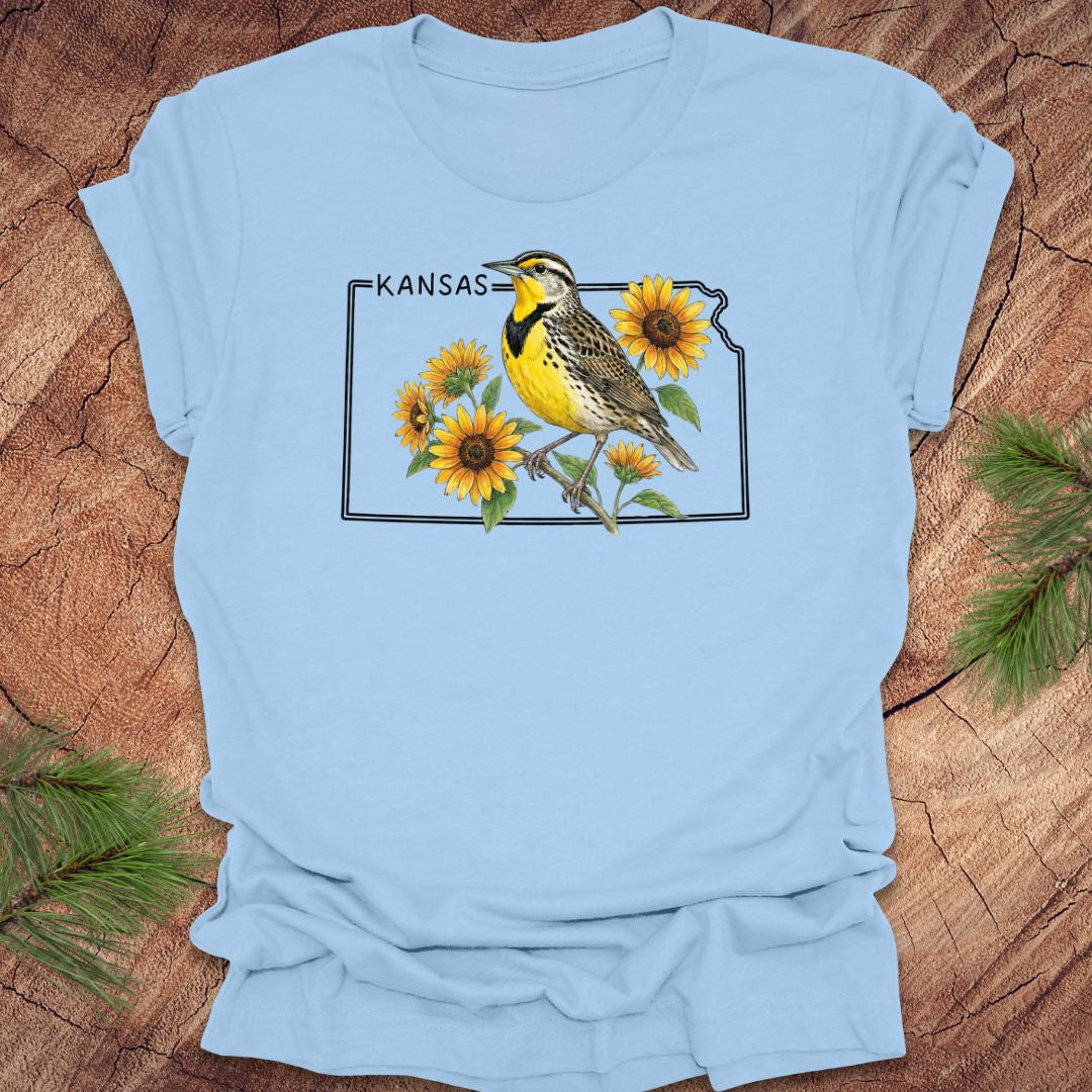 Light blue t-shirt with a graphic of a bird and sunflowers on a Kansas map, placed on a wooden surface.