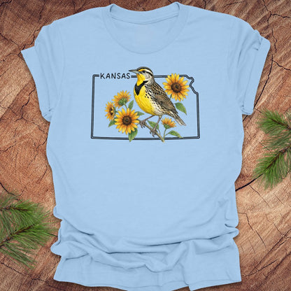 Light blue t-shirt with a graphic of a bird and sunflowers on a Kansas map, placed on a wooden surface.