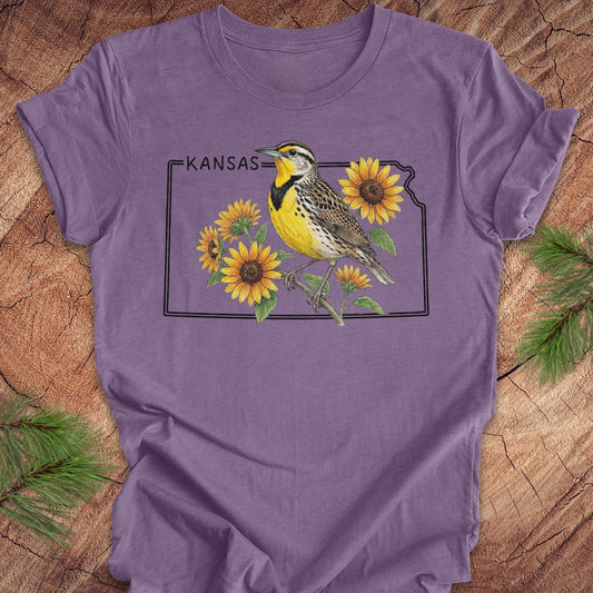 A purple short-sleeve crewneck t-shirt laid flat on a rustic wood background with pine branches. The shirt features a graphic print of a yellow and black western meadowlark bird perched among yellow sunflowers, with the word 'KANSAS' above the birds.