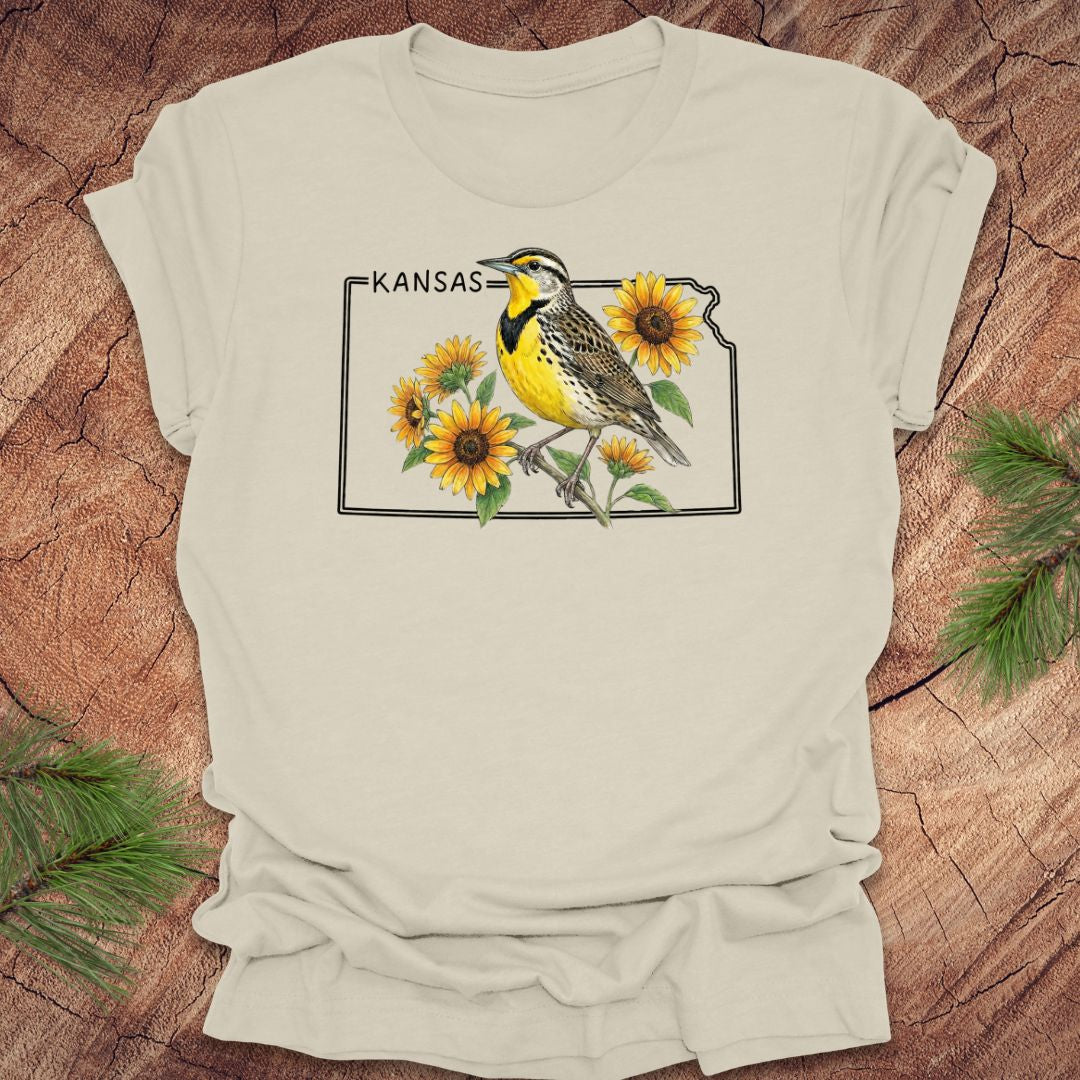 Beige t-shirt with a graphic of a bird and sunflowers on a 'Kansas' design, placed on a wooden surface.