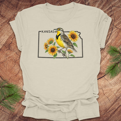 Beige t-shirt with a graphic of a bird and sunflowers on a 'Kansas' design, placed on a wooden surface.