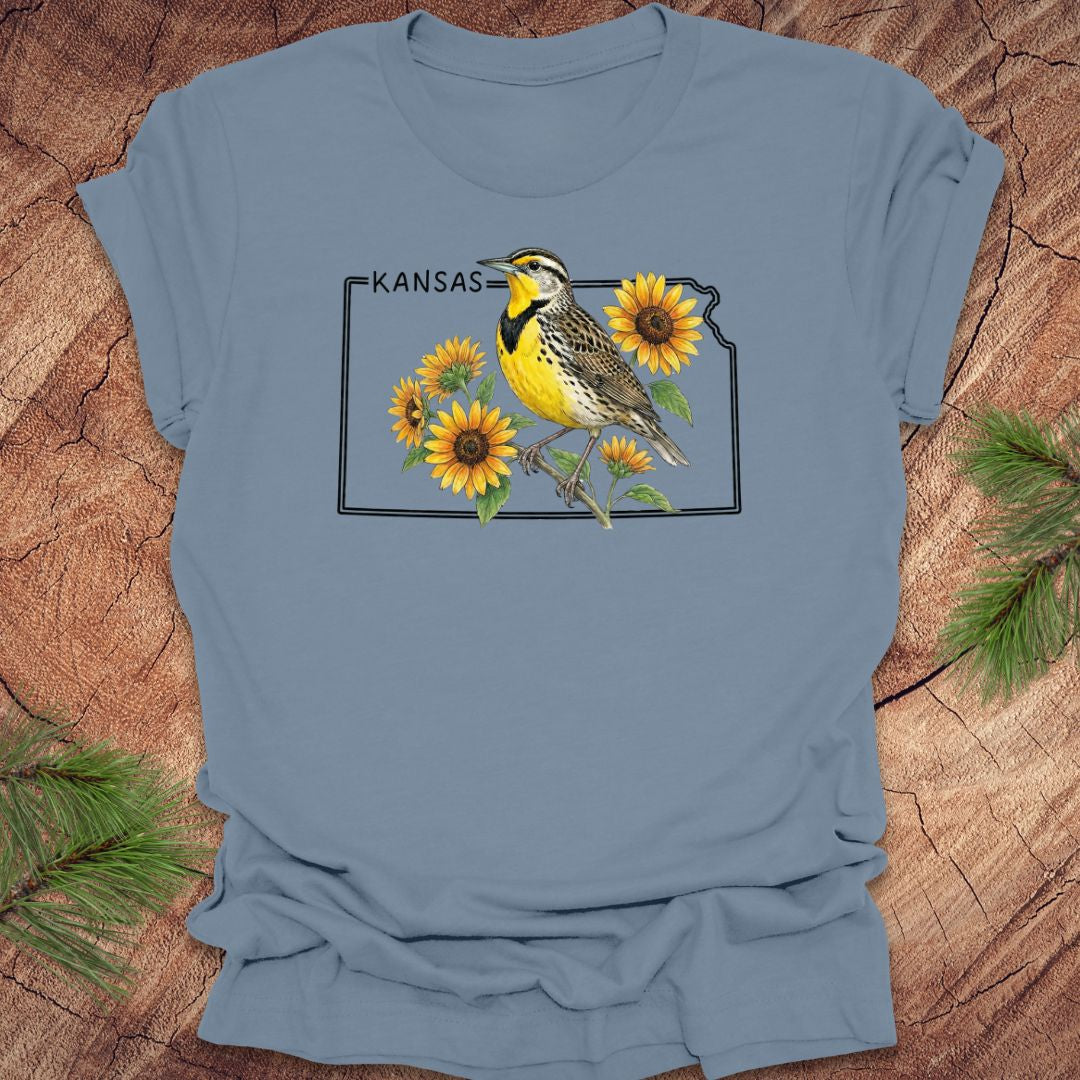 Blue t-shirt with a graphic of a bird and sunflowers on a Kansas outline design, placed on a wooden surface.