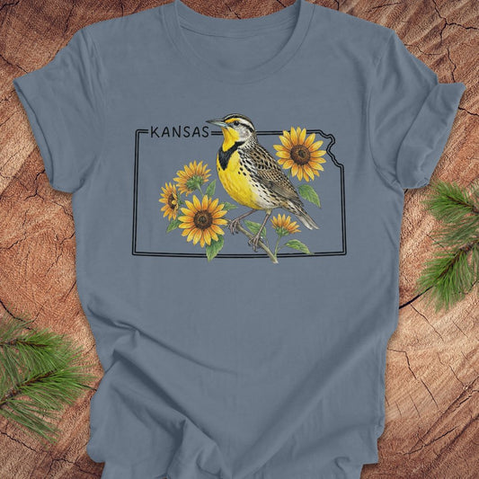 A slate blue short-sleeve t-shirt laid flat on a rustic wood background with pine branches in the corners. The shirt features a graphic print of a yellow and black bird perched among yellow sunflowers, with the word 'KANSAS' above the design.