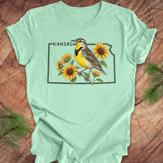Mint green t-shirt with a graphic of a bird and sunflowers on a Kansas map, placed on a wooden surface.
