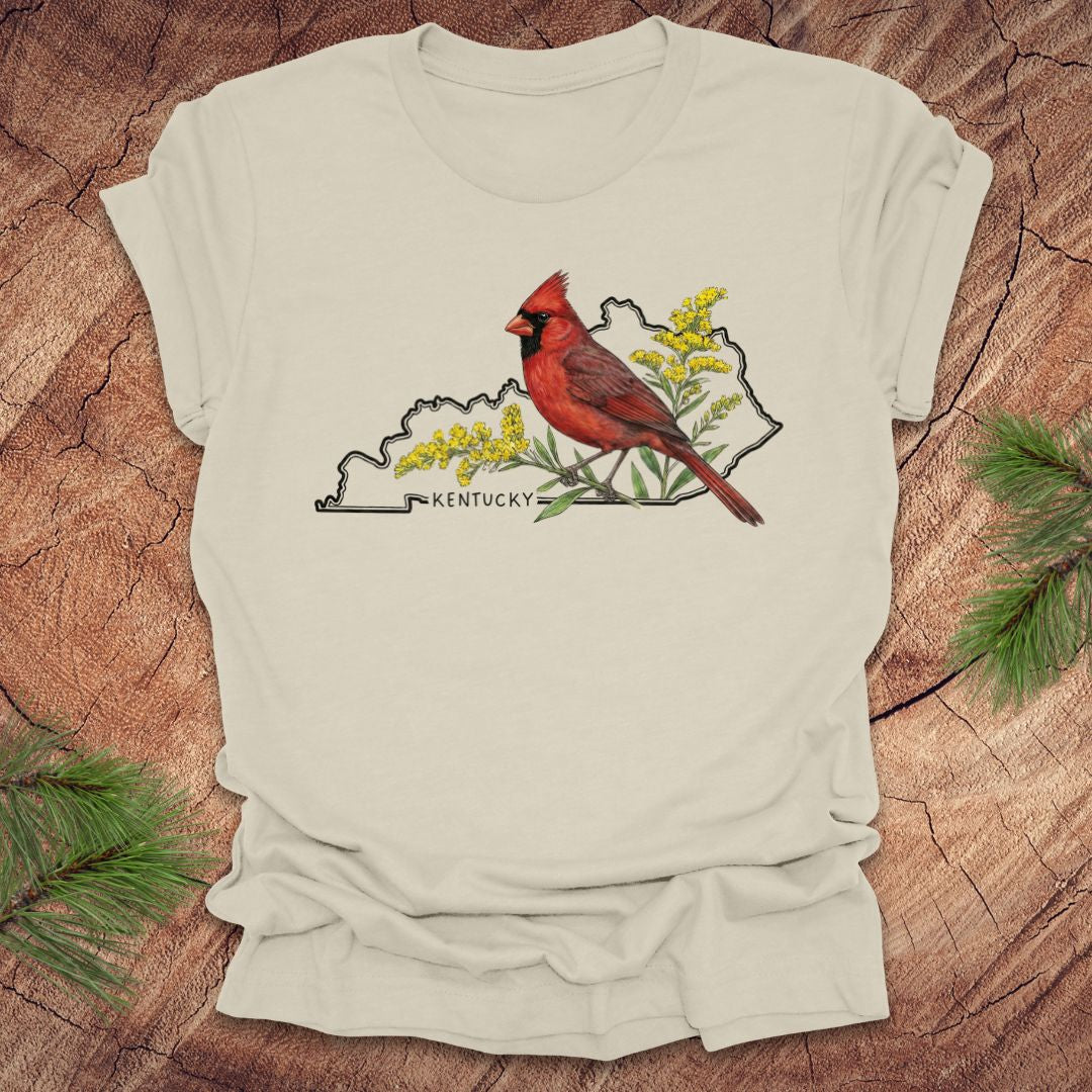 Beige t-shirt with the outline of the state of kentucky and state bird cardinal and goldenrod  design on a wooden surface