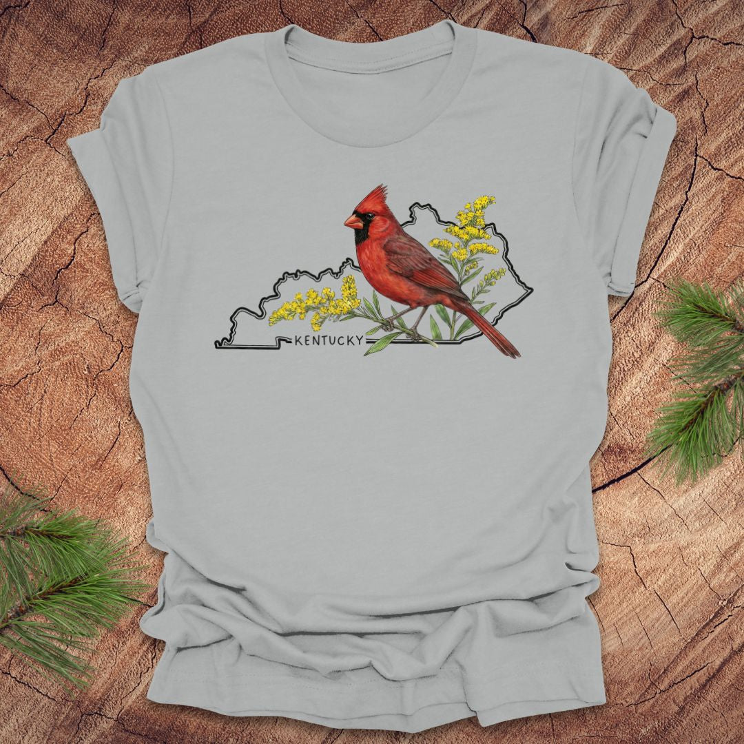 Silver t-shirt with the outline of the state of kentucky and state bird cardinal and goldenrod  design on a wooden surface