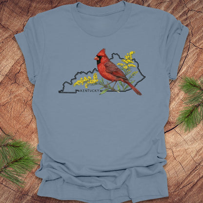 Steel blue t-shirt with the outline of the state of kentucky and state bird cardinal and goldenrod  design on a wooden surface