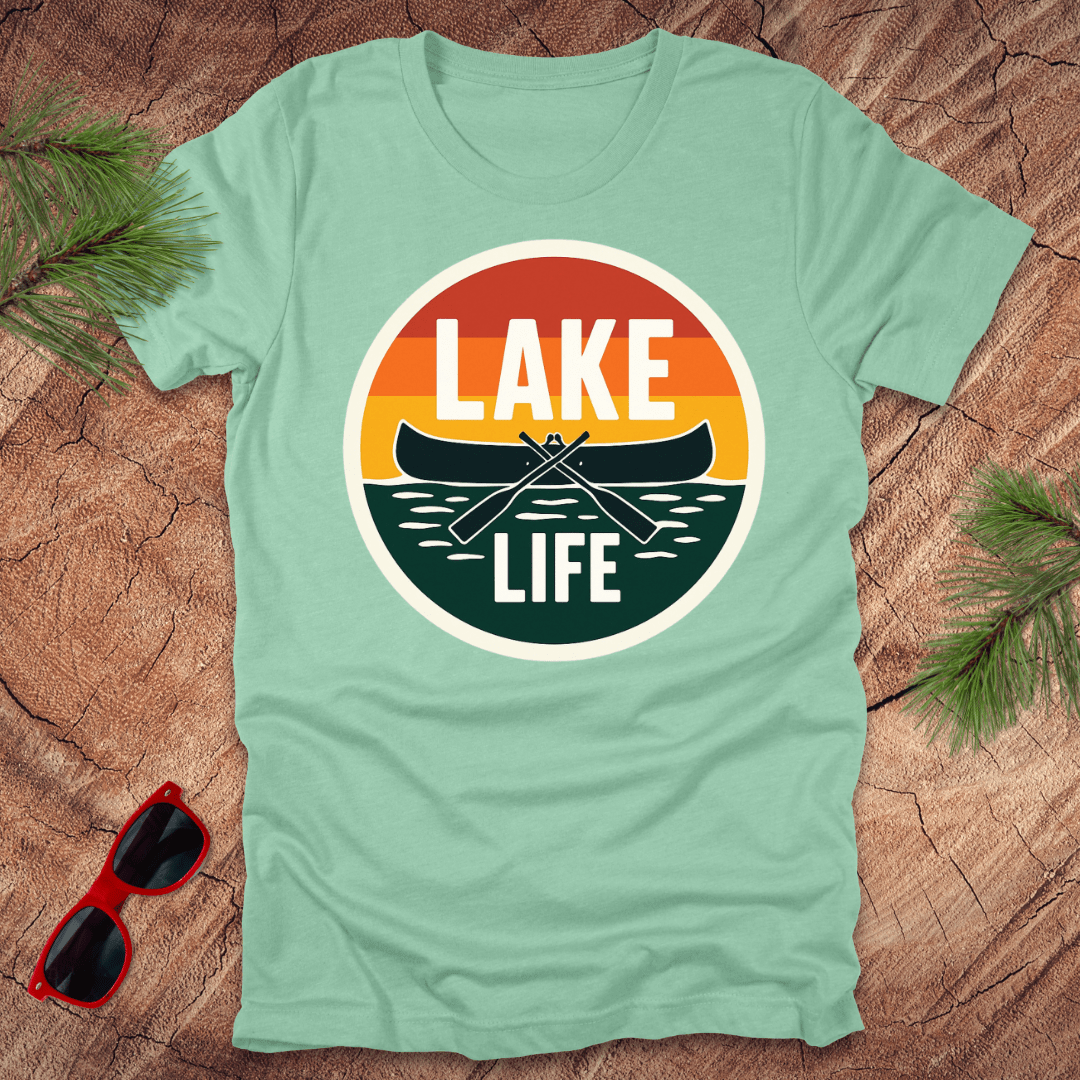 A mint green t-shirt with a vintage sunset design featuring a canoe and the text 'LAKE LIFE'. The shirt is presented on a wooden surface with pine needles and a pair of sunglasses to the side.