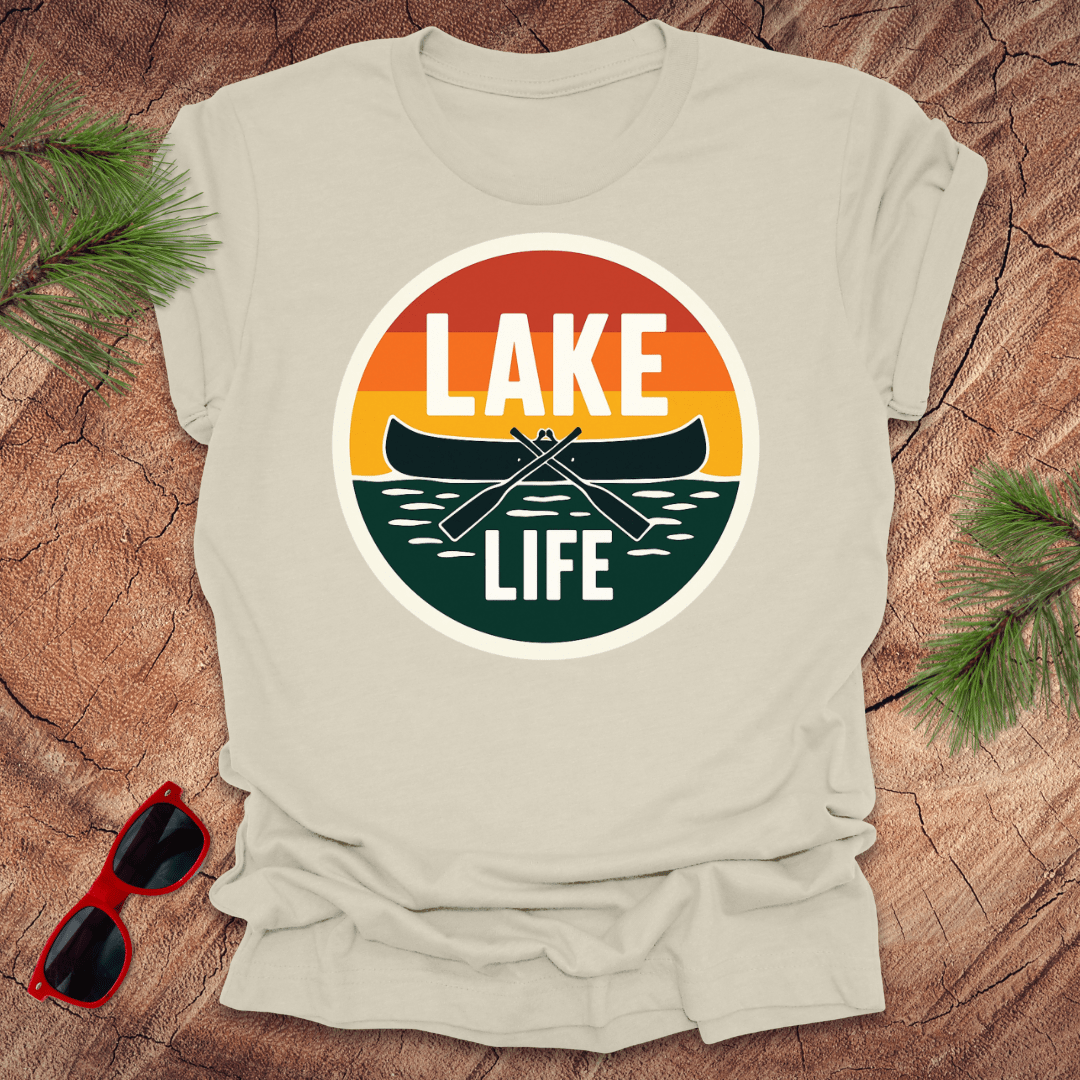 A beige t-shirt with a vintage sunset design featuring a canoe and the text 'LAKE LIFE'. The shirt is presented on a wooden surface with pine needles and a pair of sunglasses to the side.