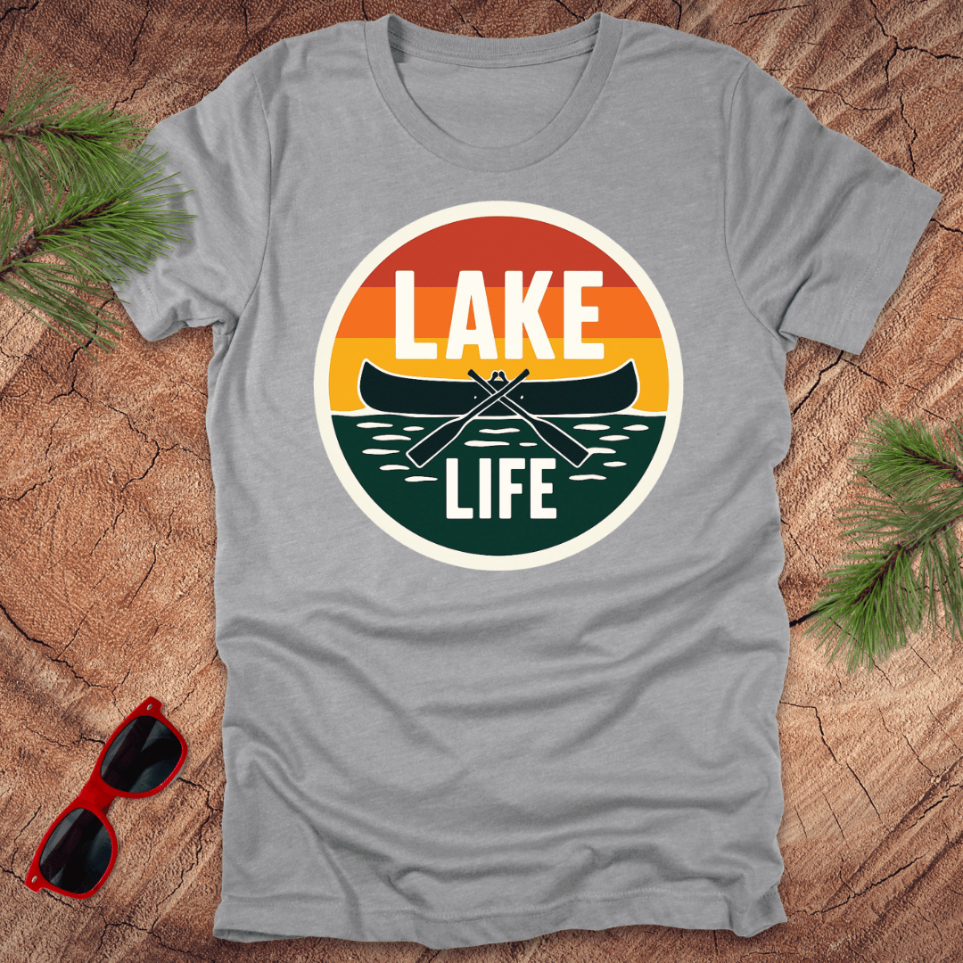 A gray t-shirt with a vintage sunset design featuring a canoe and the text 'LAKE LIFE'. The shirt is presented on a wooden surface with pine needles and a pair of sunglasses to the side.