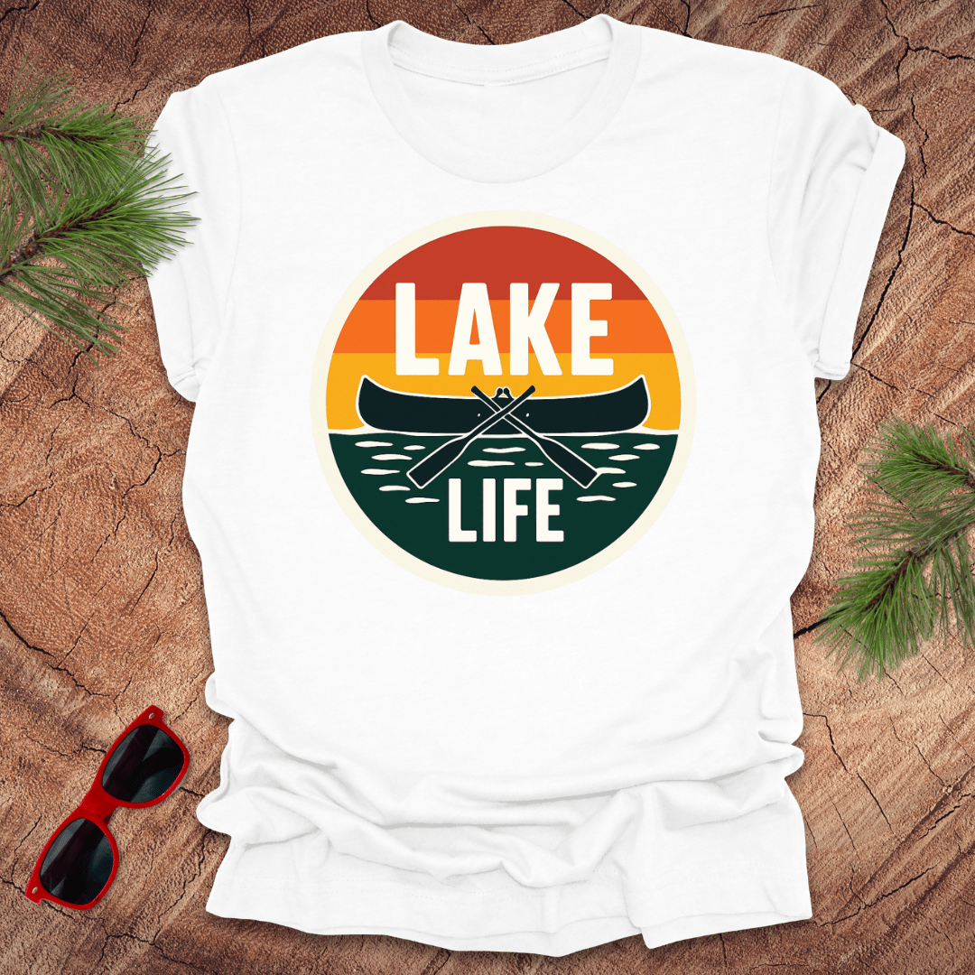 A white t-shirt with a vintage sunset design featuring a canoe and the text 'LAKE LIFE'. The shirt is presented on a wooden surface with pine needles and a pair of sunglasses to the side.