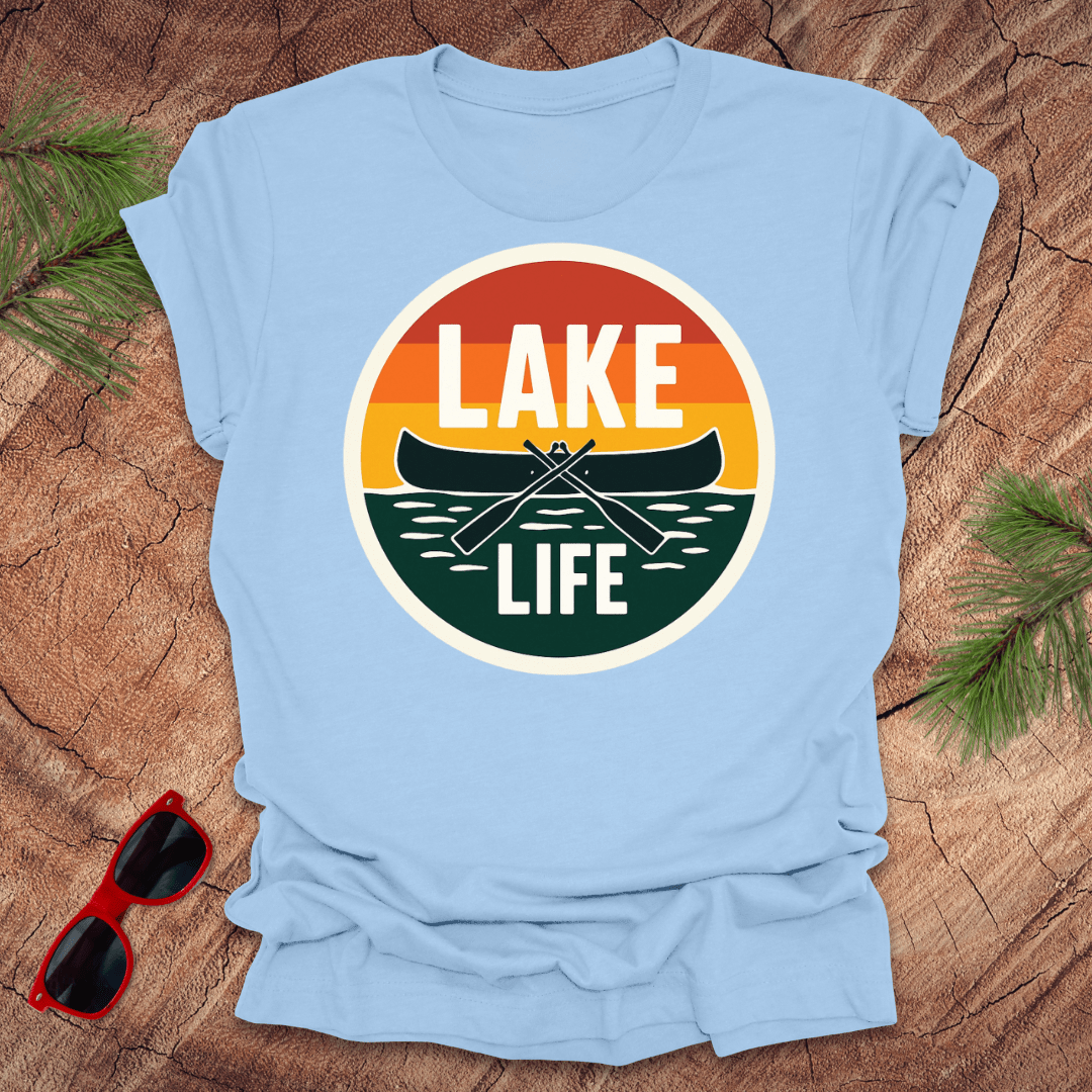 A blue t-shirt with a vintage sunset design featuring a canoe and the text 'LAKE LIFE'. The shirt is presented on a wooden surface with pine needles and a pair of sunglasses to the side.