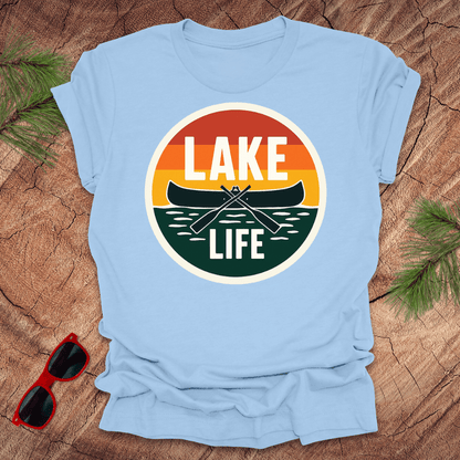 A blue t-shirt with a vintage sunset design featuring a canoe and the text 'LAKE LIFE'. The shirt is presented on a wooden surface with pine needles and a pair of sunglasses to the side.