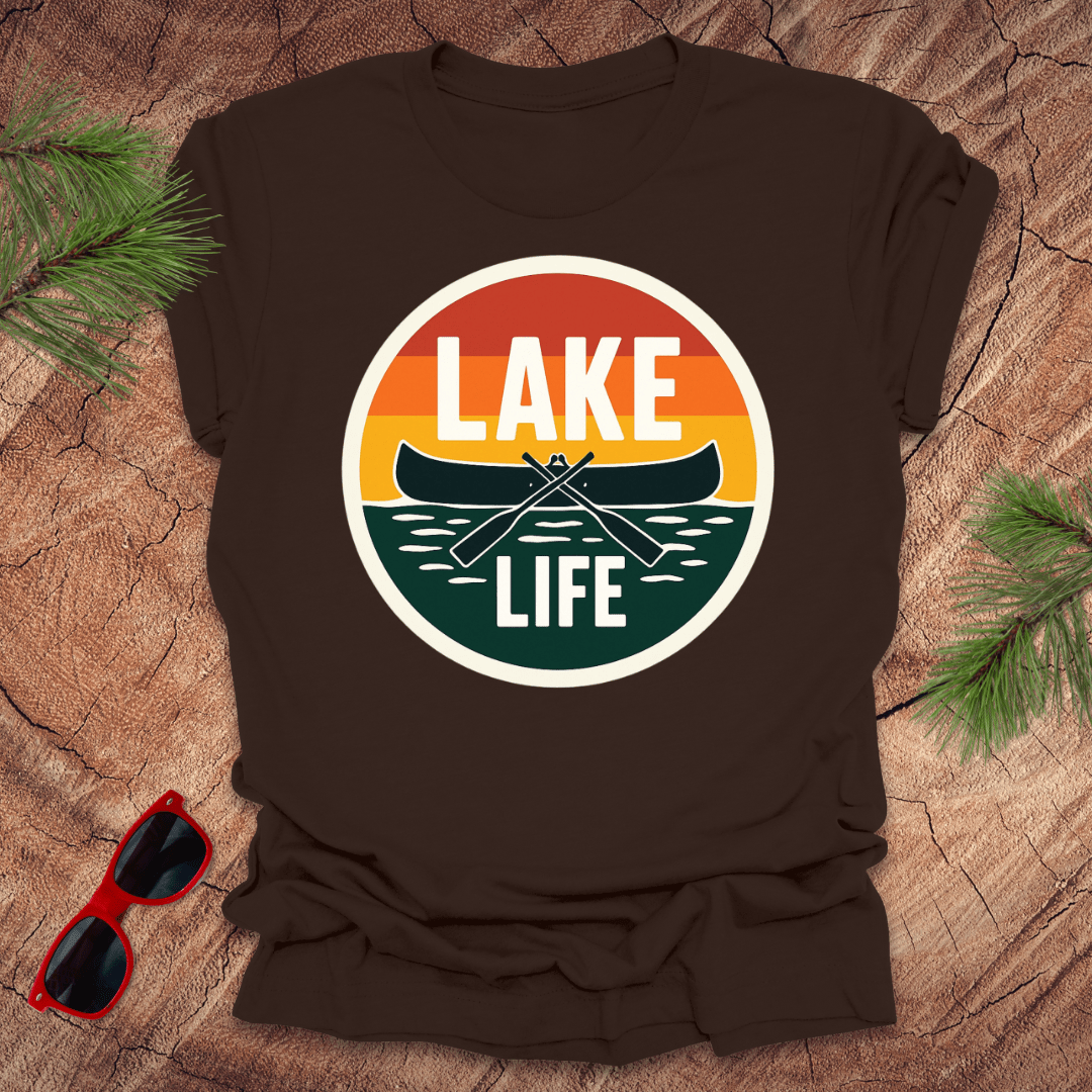 A brown t-shirt with a vintage sunset design featuring a canoe and the text 'LAKE LIFE'. The shirt is presented on a wooden surface with pine needles and a pair of sunglasses to the side.