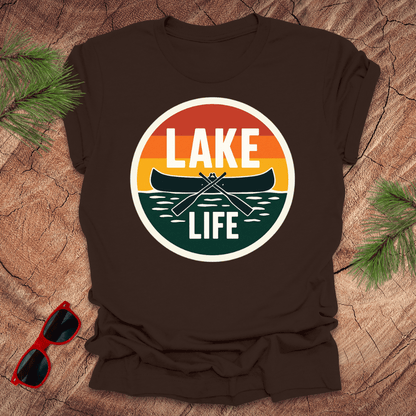 A brown t-shirt with a vintage sunset design featuring a canoe and the text 'LAKE LIFE'. The shirt is presented on a wooden surface with pine needles and a pair of sunglasses to the side.