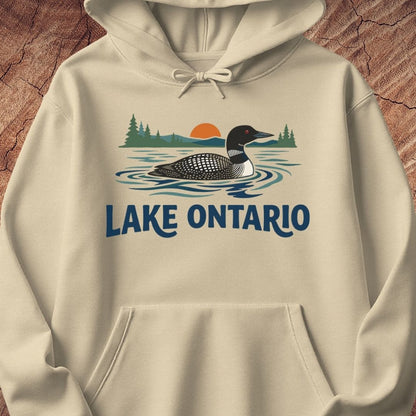 A cream-colored pullover hoodie featuring a graphic print of a loon on Lake Ontario with pine trees and a sunset, displayed on a textured wood background.
