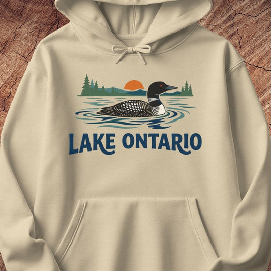 A cream-colored pullover hoodie featuring a graphic print of a loon on Lake Ontario with pine trees and a sunset, displayed on a textured wood background.