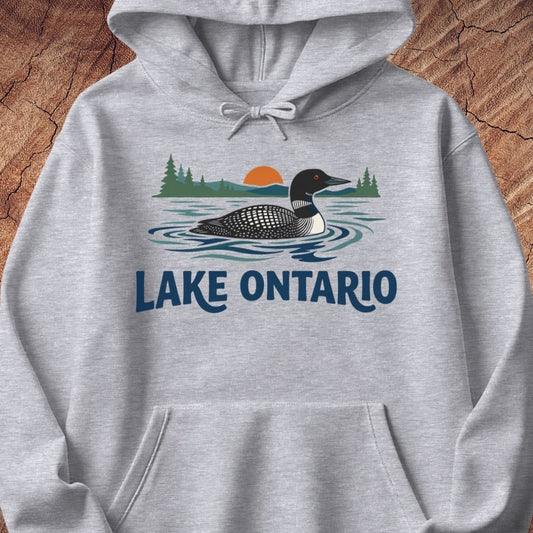 A light heather gray pullover hoodie featuring a graphic print of a loon duck on Lake Ontario with pine trees, a sunset, and the text 'LAKE ONTARIO' in dark blue. The hoodie is displayed flat against a rustic wood background.