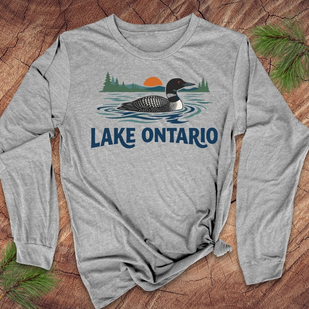 A heather gray long-sleeve t-shirt with a graphic of a loon duck on Lake Ontario, featuring pine trees and an orange sun, laid flat on a rustic wood background with pine needles.