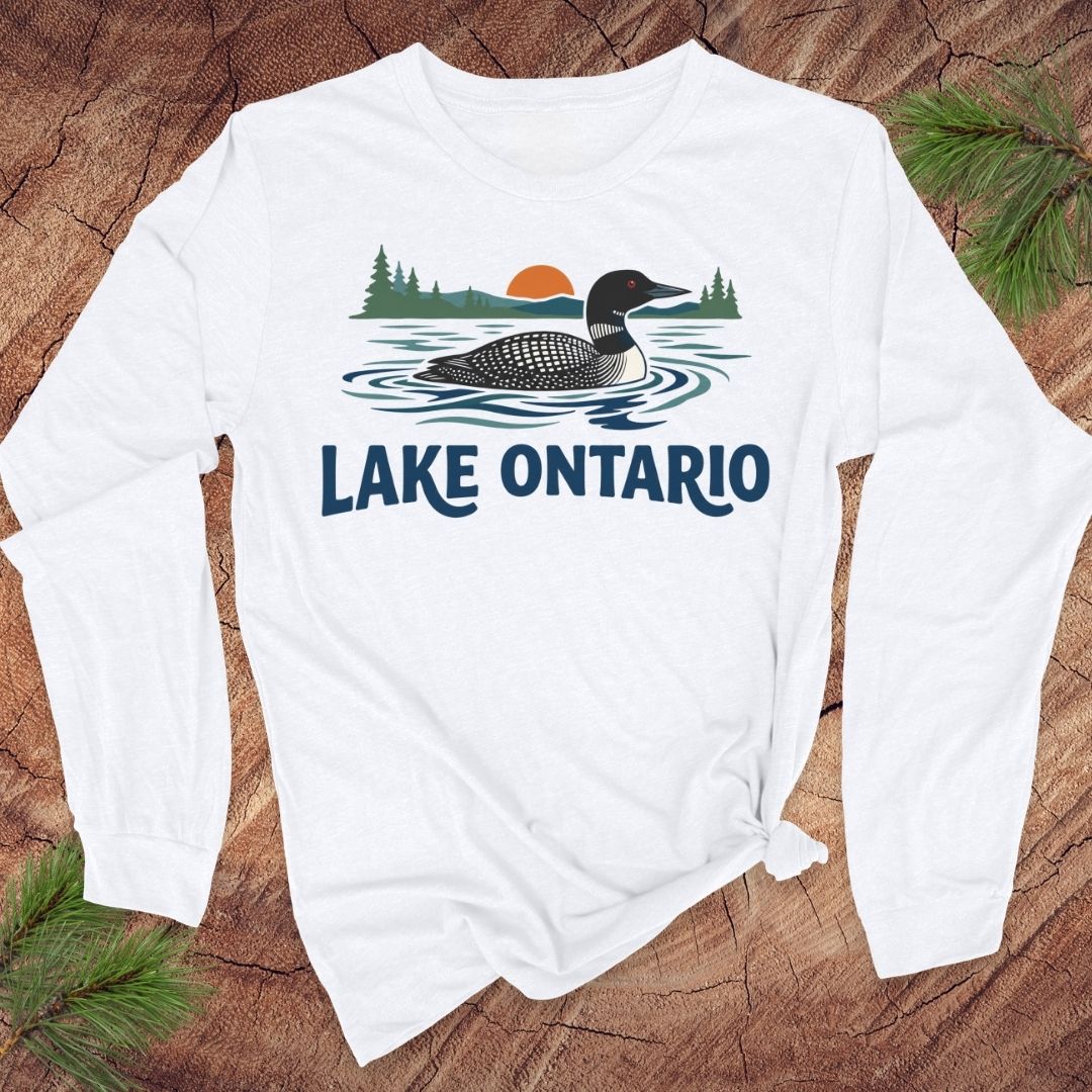 A white long-sleeve t-shirt with a 'Lake Ontario' loon graphic, styled flat on a rustic wood background with pine branches.