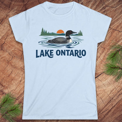 A light blue short-sleeve t-shirt with a graphic of a loon on Lake Ontario, featuring pine trees, a sunset, and the text 'LAKE ONTARIO' in navy blue. The t-shirt is displayed flat on a rustic wood background with pine needles in the corners.