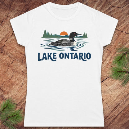 A white short-sleeve crewneck t-shirt featuring a graphic of a loon on Lake Ontario with pine trees and a sunset, displayed flat on a rustic wood background with pine needles.