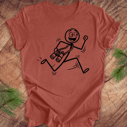 Clay colored t-shirt with a stick figure design running with binoculars on a wooden surface