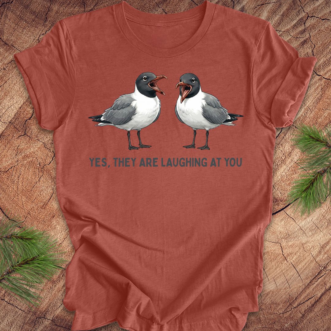 Clay colored t-shirt with two laughing gulls and text, "yes, they are laughing at you" on a wood background.