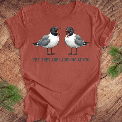 Clay colored t-shirt with two laughing gulls and text, "yes, they are laughing at you" on a wood background.